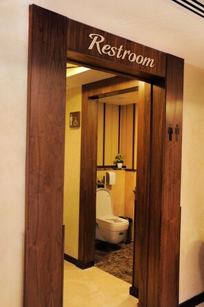 Una porta in legno con la scritta 'Restroom' che dà accesso a un bagno con un gabinetto, un asciugamani ad aria e una pianta in vaso.
