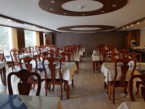 Zona pranzo con tavoli e sedie, soffitto con decorazioni circolari e vista su alberi attraverso ampie finestre.