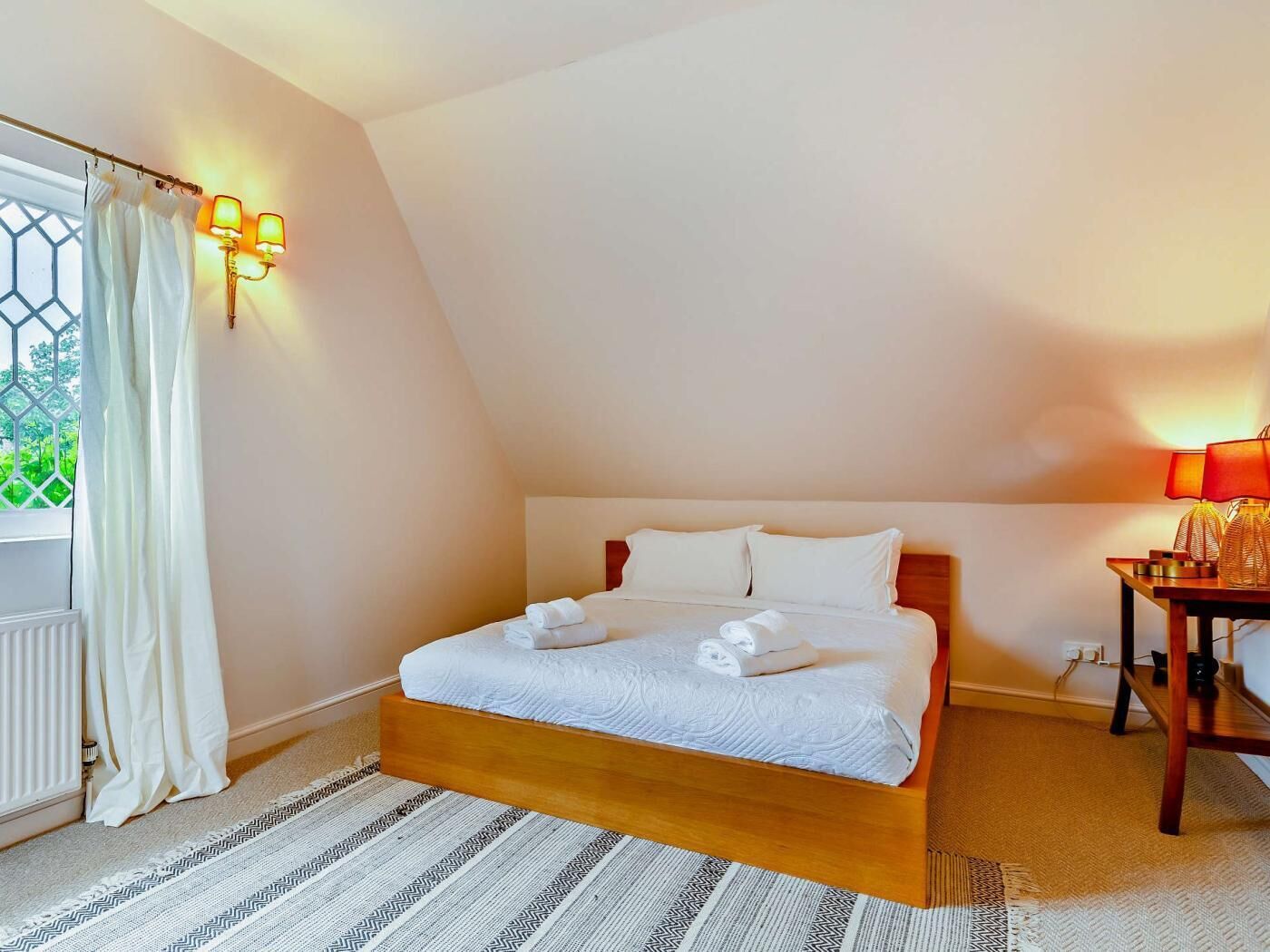 A bedroom with a bed, a window with curtains, a bedside table with a lamp, and a radiator.