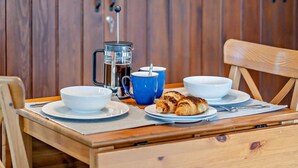 Una mesa de madera con una cafetera, tazas azules y blancas, un plato con cruasanes y una silla.