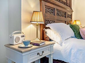 Een opgemaakt bed met witte lakens, een bedlampje, een radio, een boek en een beker op een nachtkastje.