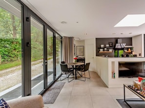 A modern interior with a dining area, kitchen, and large sliding glass doors offering a view of a forested path.
