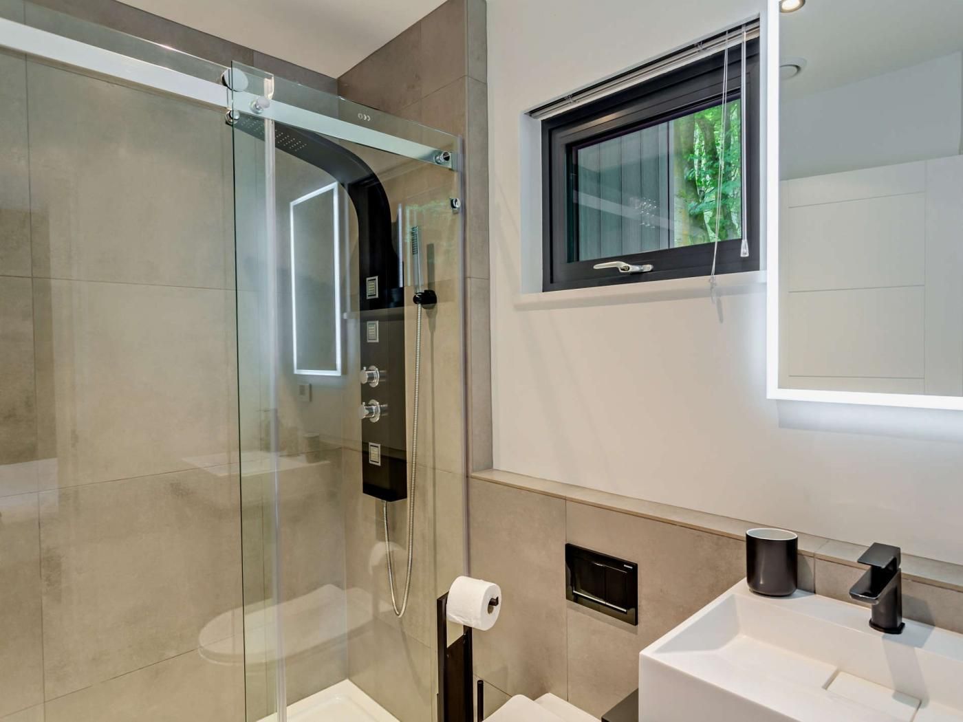 A modern bathroom with a glass-enclosed shower, a toilet paper holder, a window with a view, and a sink with a faucet.
