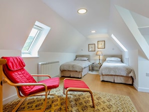 A room with two beds, a red chair, a small table with a lamp, and a radiator.