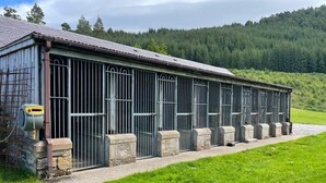 Ein längliches Gebäude mit vergitterten Fenstern und einem Steinfundament, umgeben von einer grünen Fläche und einem bewaldeten Hügel im Hintergrund.
