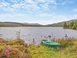 A calm lake with a green boat docked on the shore, surrounded by grassy areas and hills in the background. - 1 Bed in Ardross (oc-95533) (Alness)