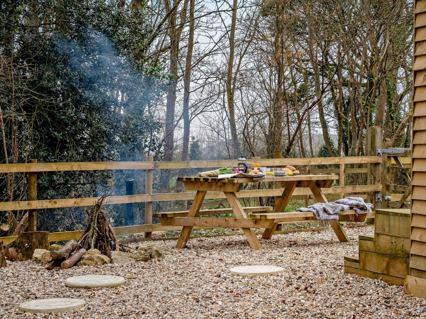 A wooden picnic table with food items, surrounded by a wooden fence and trees.
