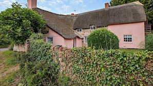 Ein traditionelles Haus mit Strohdach und rosa Wänden, umgeben von viel Grün und einer Steinmauer.