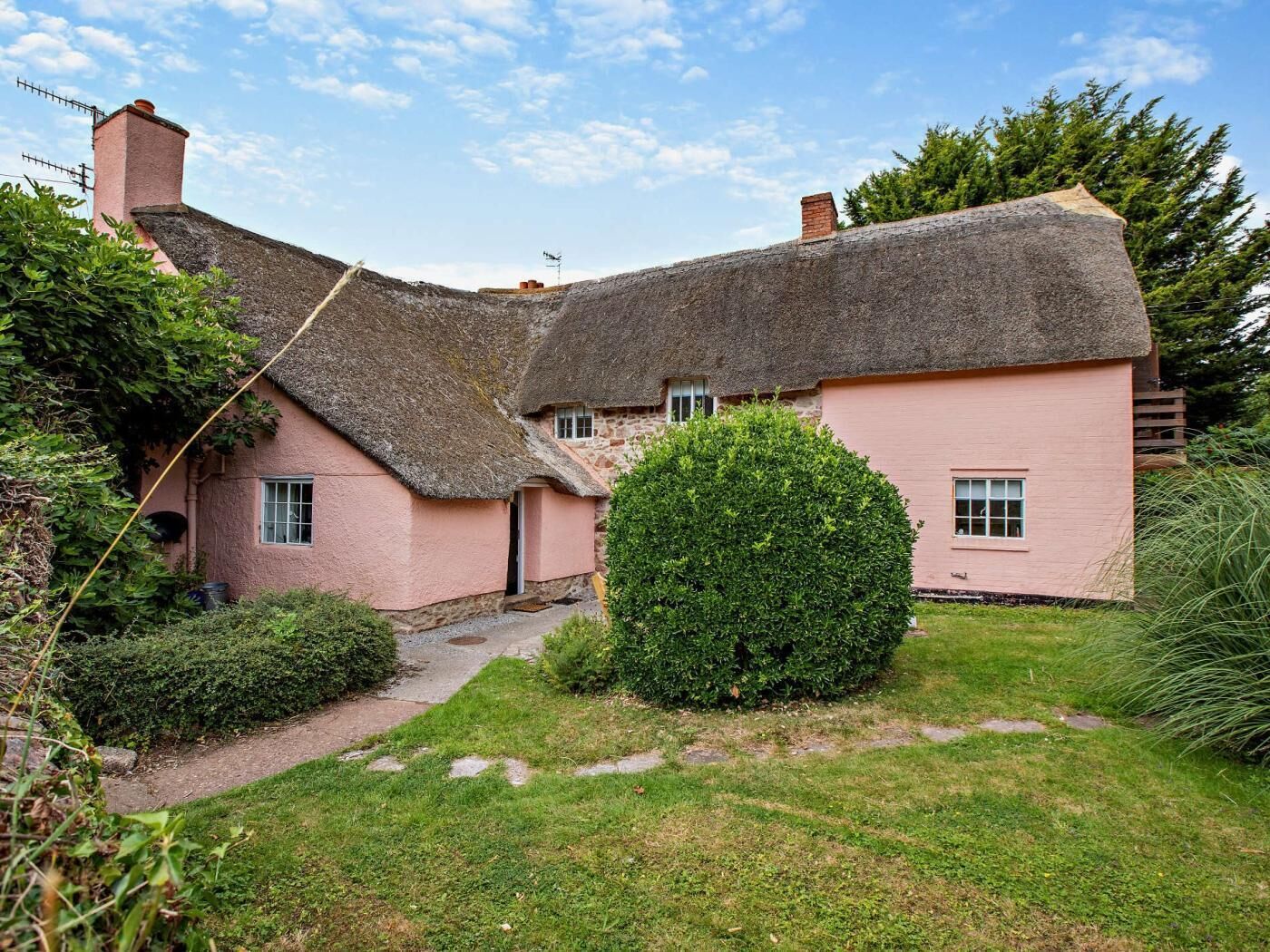 A traditional thatched-roof cottage with a pink exterior, a stone chimney, and a well-maintained garden with a hedge and a small bush.