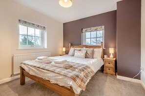 Un dormitorio con una cama de madera, dos mesitas de noche con lámparas, un radiador y ventanas con cortinas.