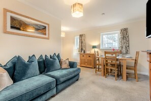 A living room with a sofa, a dining table, and chairs. There is a painting on the wall, a lamp, and a window with curtains. - 3 Bed in Workington (oc-92628) (Dean)