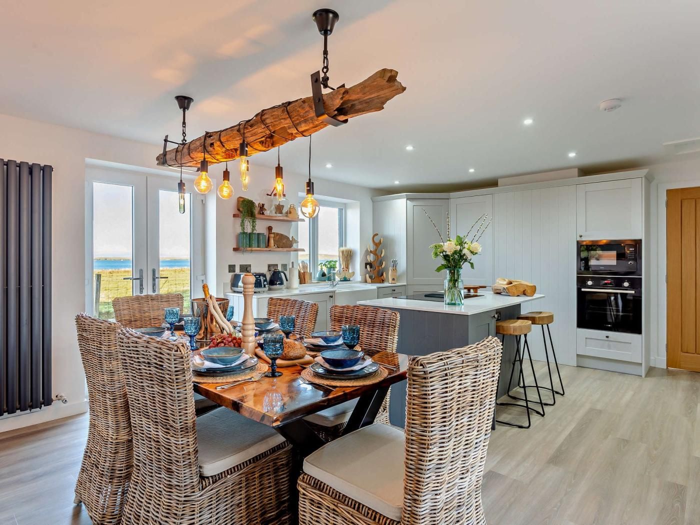 Une cuisine moderne avec des meubles blancs, un îlot central et un coin repas agrémenté de chaises en osier et d’une table en bois mise pour le repas.