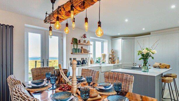 Cocina moderna con comedor, con una mesa de madera dispuesta para una comida, sillas de mimbre y vista al mar.