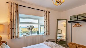 Un dormitorio con cama, ventana con cortinas, radiador, una silla, un espejo y una lámpara colgante.