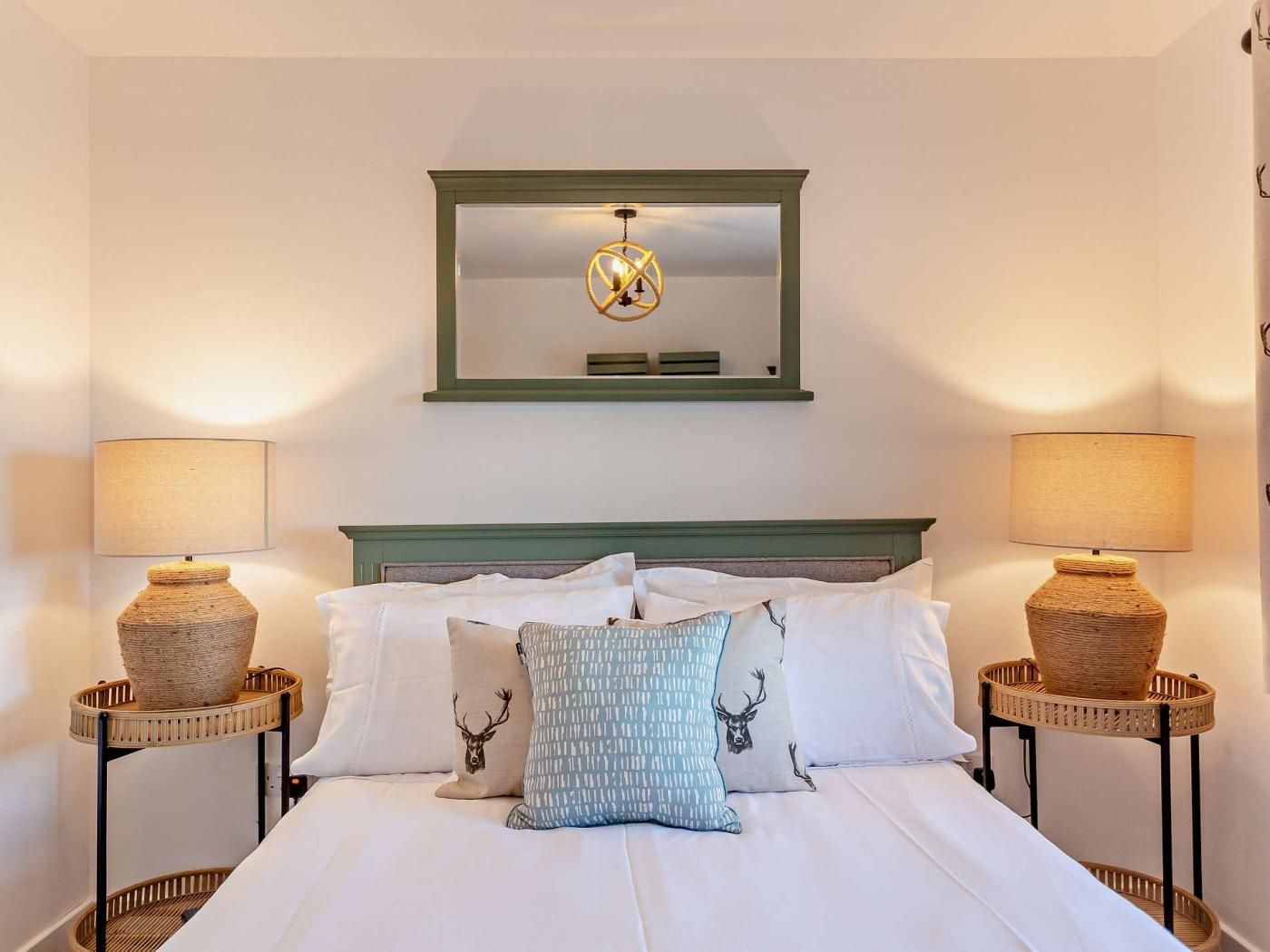 Ein ordentlich bezogenes Bett mit weißen Leinentextilien und dekorativen Kissen, begleitet von zwei Nachttischen mit gewebten Körben, einem spiegelnden Wandbild in grünem Rahmen und einer an der Wand befestigten Lampe.