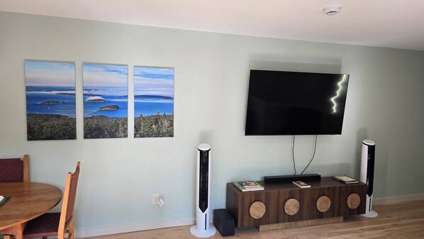 A living room with a flat-screen TV mounted on the wall, two tall floor-standing speakers, a wooden dining table with chairs, and two landscape paintings on the wall.