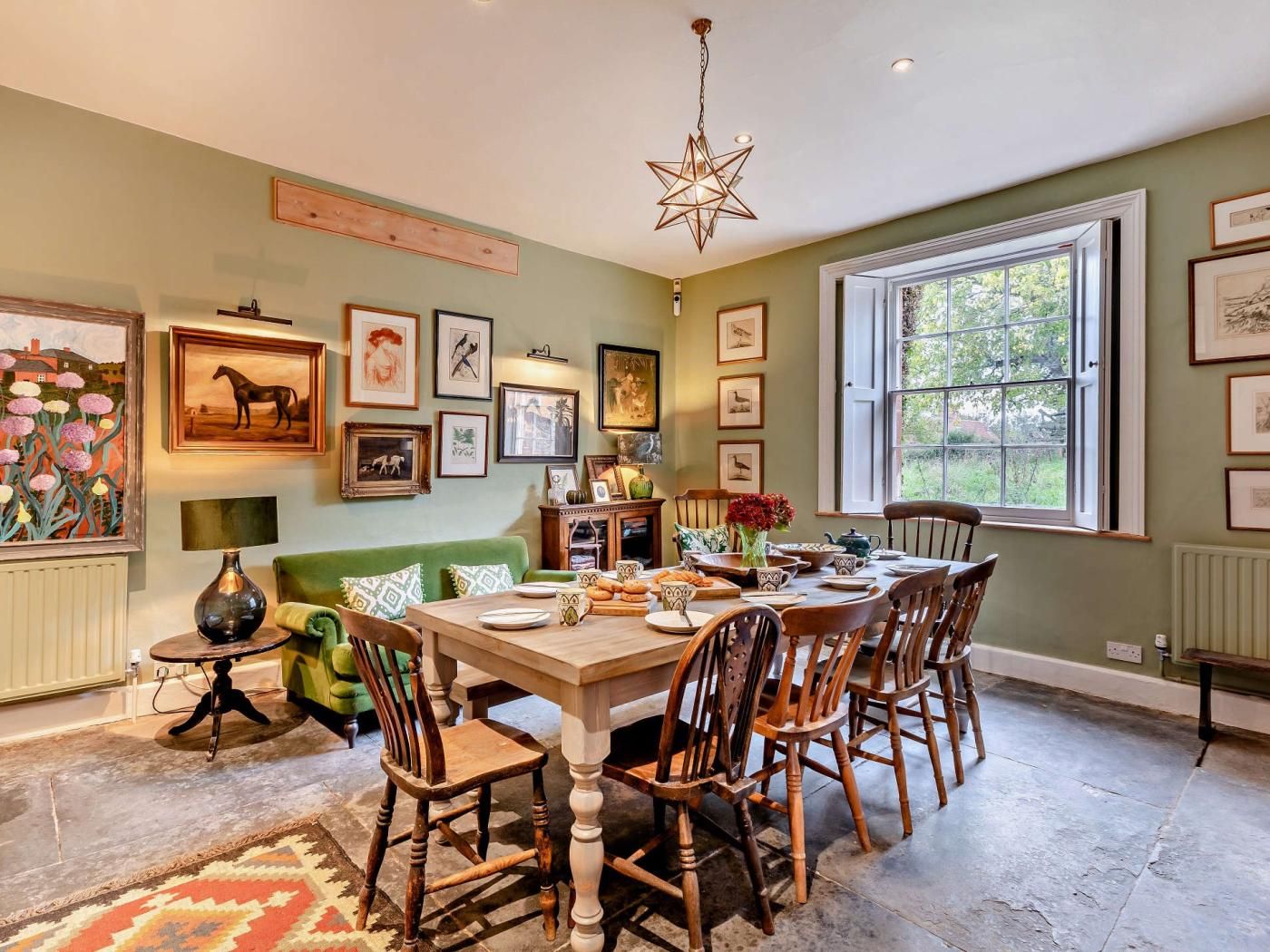 A dining area with a wooden table, chairs, and a chandelier. There are framed pictures on the walls and a window with a view of greenery.
