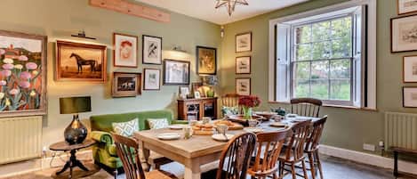 A dining area with a wooden table, chairs, and a chandelier. There are framed pictures on the walls and a window with a view of greenery.