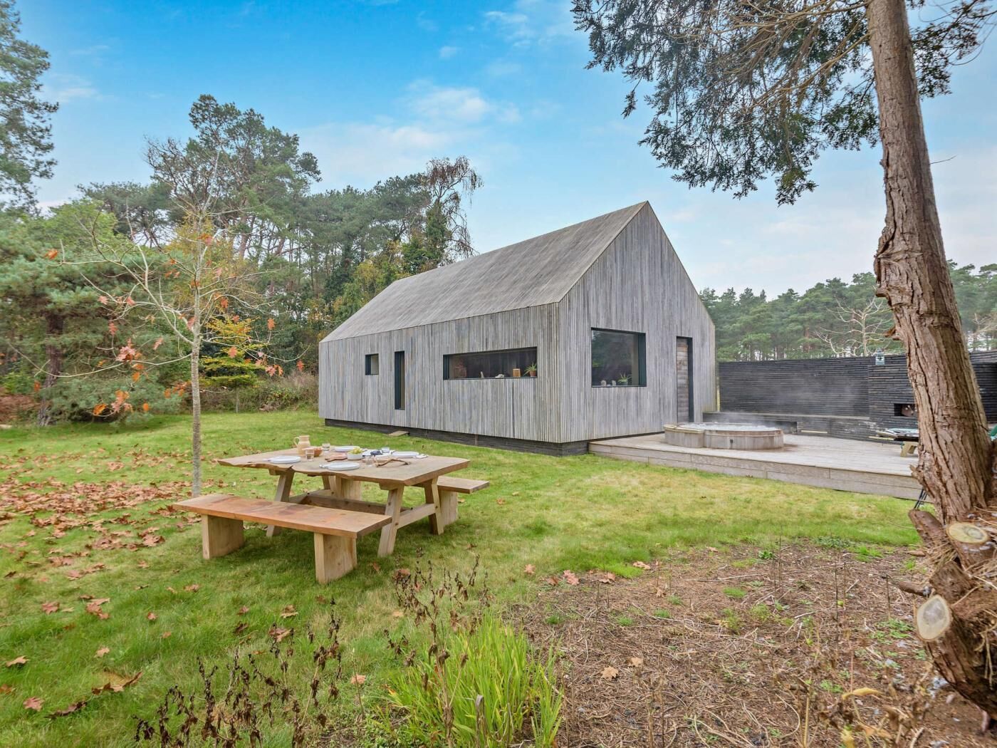 A modern, wooden cabin with a sloped roof, a patio with a table and benches, and a surrounding grassy area with trees.