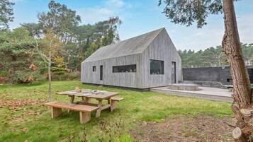 A modern, wooden cabin with a sloped roof, a patio with a table and benches, and a surrounding grassy area with trees.