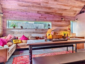 Ein gemütliches Wohnzimmer mit Holzbalkendecke, einem großen Fenster mit Blick, einer Couch mit bunten Kissen, einem Couchtisch und einem Schreibtisch mit einem Laptop.