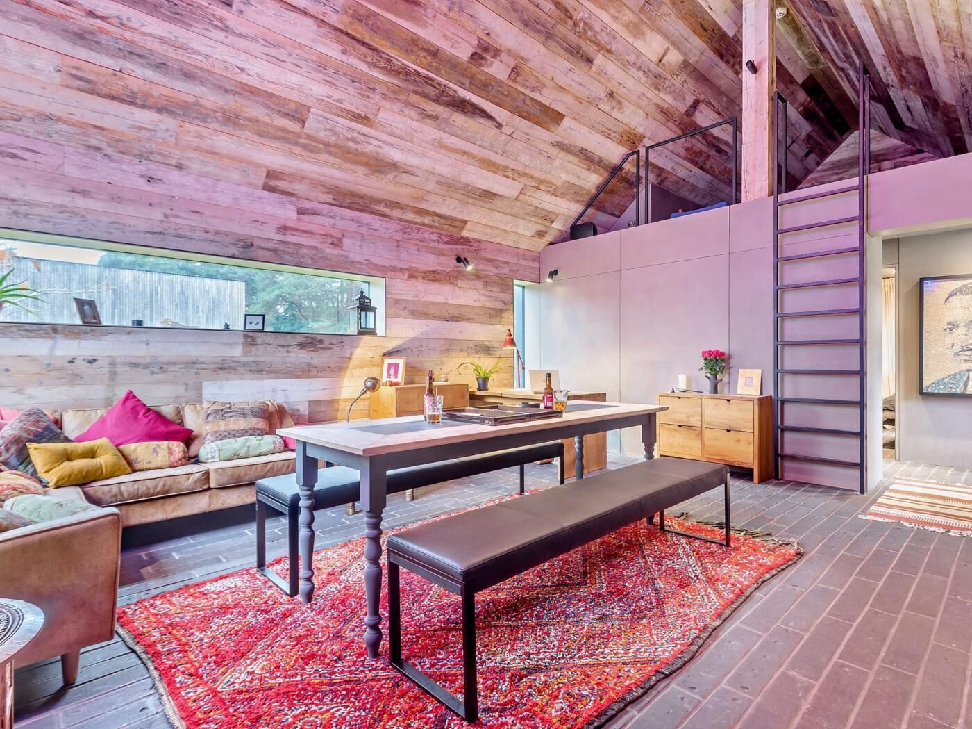 A modern interior with a wooden ceiling, a dining area with a bench, a sofa with cushions, and a small table with a bottle.