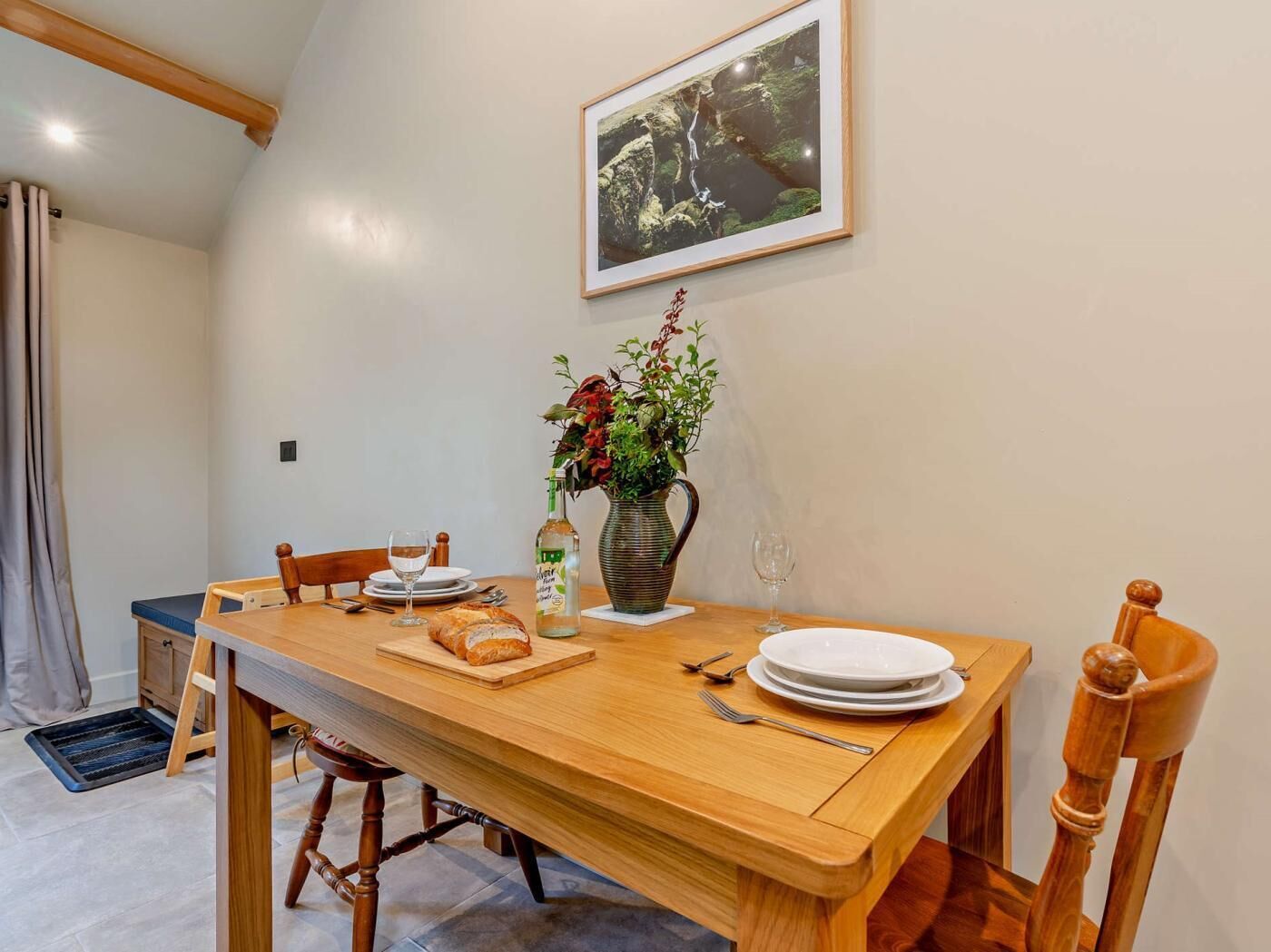 Un comedor con una mesa de madera, sillas, un florero con flores, una botella y un cuadro en la pared.
