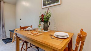 Uma sala de jantar com uma mesa de madeira, cadeiras, um vaso com flores, uma garrafa e um quadro na parede.