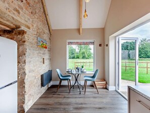 A kitchen with a brick wall, a dining table with chairs, a window with a view of a garden, and a hanging light fixture. - 1 Bed in Wedmore (oc-95582) (Theale)