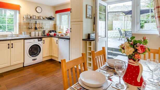 Een compacte keuken met een wasmachine, een eethoek met een tafel voor twee, en uitzicht op de tuin door de openslaande deur.