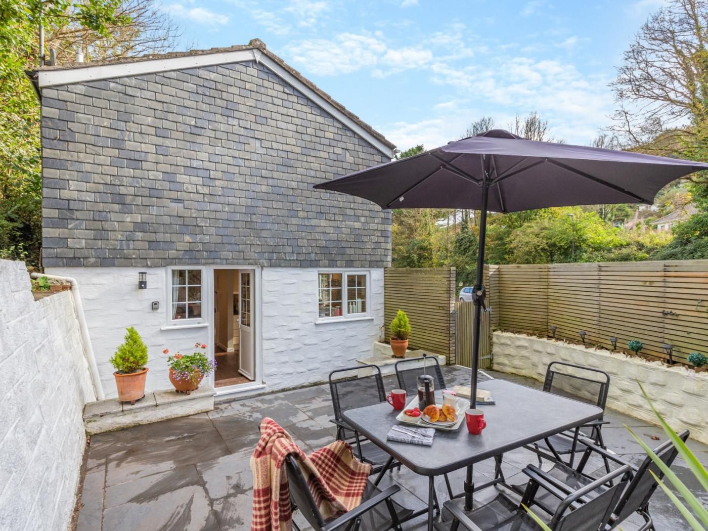 A patio area with a table, chairs, and an umbrella. There are potted plants and a small building in the background.