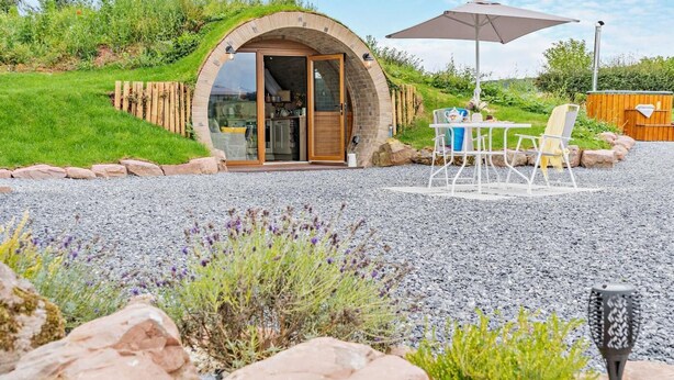 Ein rundes Steinhaus mit Glastür, Sitzgelegenheiten draußen und einem Sonnenschirm auf der Terrasse.