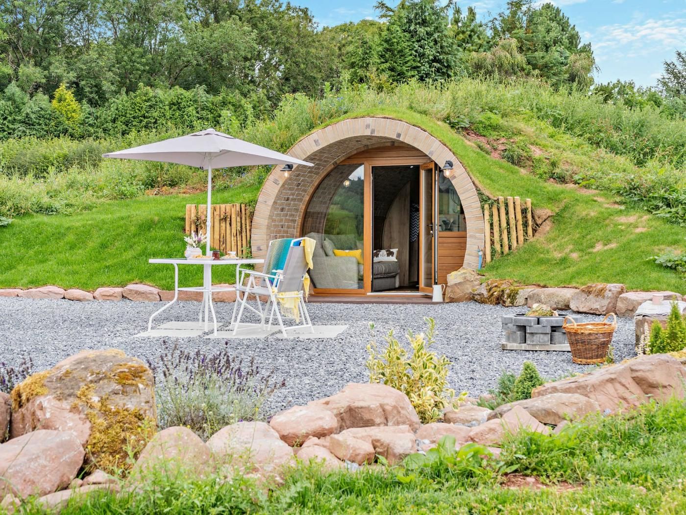 Une structure accueillante et semi-ouverte, dotée d’un toit incurvé, d’un espace salon avec une table et des chaises, et d’un petit jardin agrémenté de rochers et de plantes.