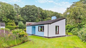 A small, single-story house with a blue door and white walls, surrounded by a well-maintained garden and dense forest. - 2 Bed in Saltash (oc-95642) (Notter)