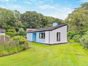 Ein kleines, einstöckiges Haus mit blauer Tür und weißen Wänden, umgeben von einem gepflegten Garten und dichtem Wald.