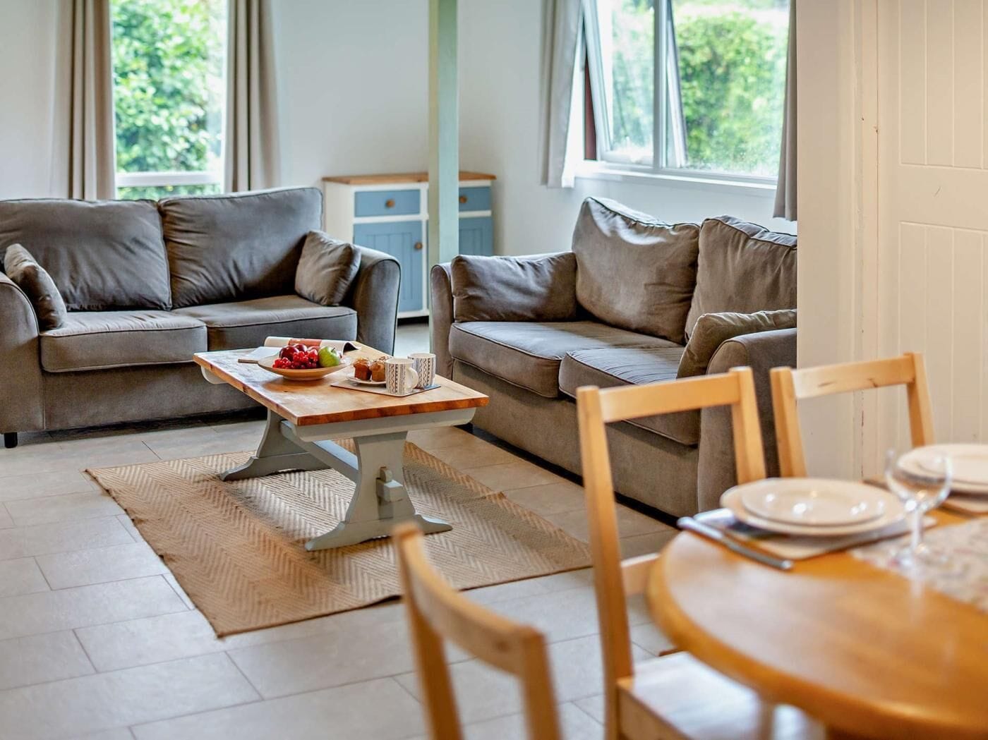 Un salón con dos sofás, una mesa de centro con frutas y tazas, una mesa de comedor con platos y vasos, y una ventana con vistas a zonas verdes.