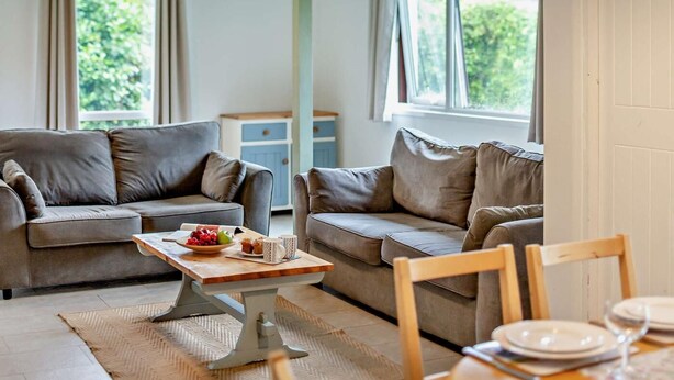 A living room with two sofas, a coffee table with fruits and mugs, a dining table with plates and glasses, and a window with a view of greenery. - 2 Bed in Saltash (oc-95642) (Notter)