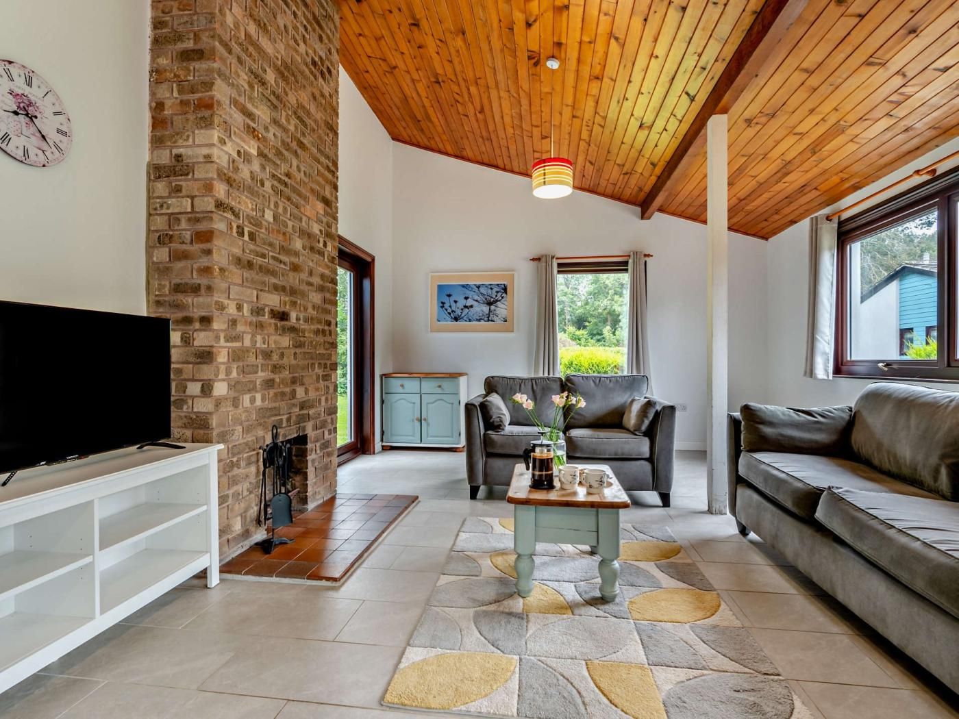 A living room with a brick fireplace, a television, a sofa, a coffee table, and a view of greenery outside.