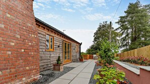 A brick building with a wooden door, a paved patio, and a garden with plants and a large pot. - 1 Bed in Bromyard (oc-95609) (Pencombe)