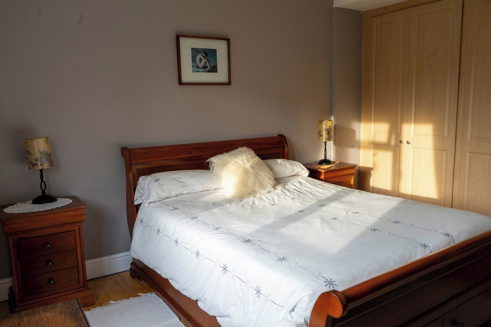 A bedroom with a wooden bed, two bedside tables, a nightstand, a framed picture on the wall, and a wardrobe.