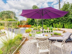 Een terras buiten met een ronde tafel, witte stoelen, een paarse parasol en een tuin met verschillende planten en een houten hek.