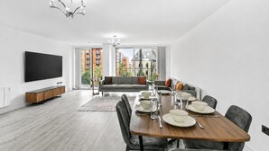 Un salon moderne comprenant un coin repas, un téléviseur à écran plat et une vue sur les immeubles grâce à de grandes fenêtres.