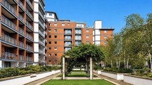 Un immeuble résidentiel à plusieurs étages, doté de balcons, d’une cour intérieure agrémentée d’une pergola et entouré de verdure.