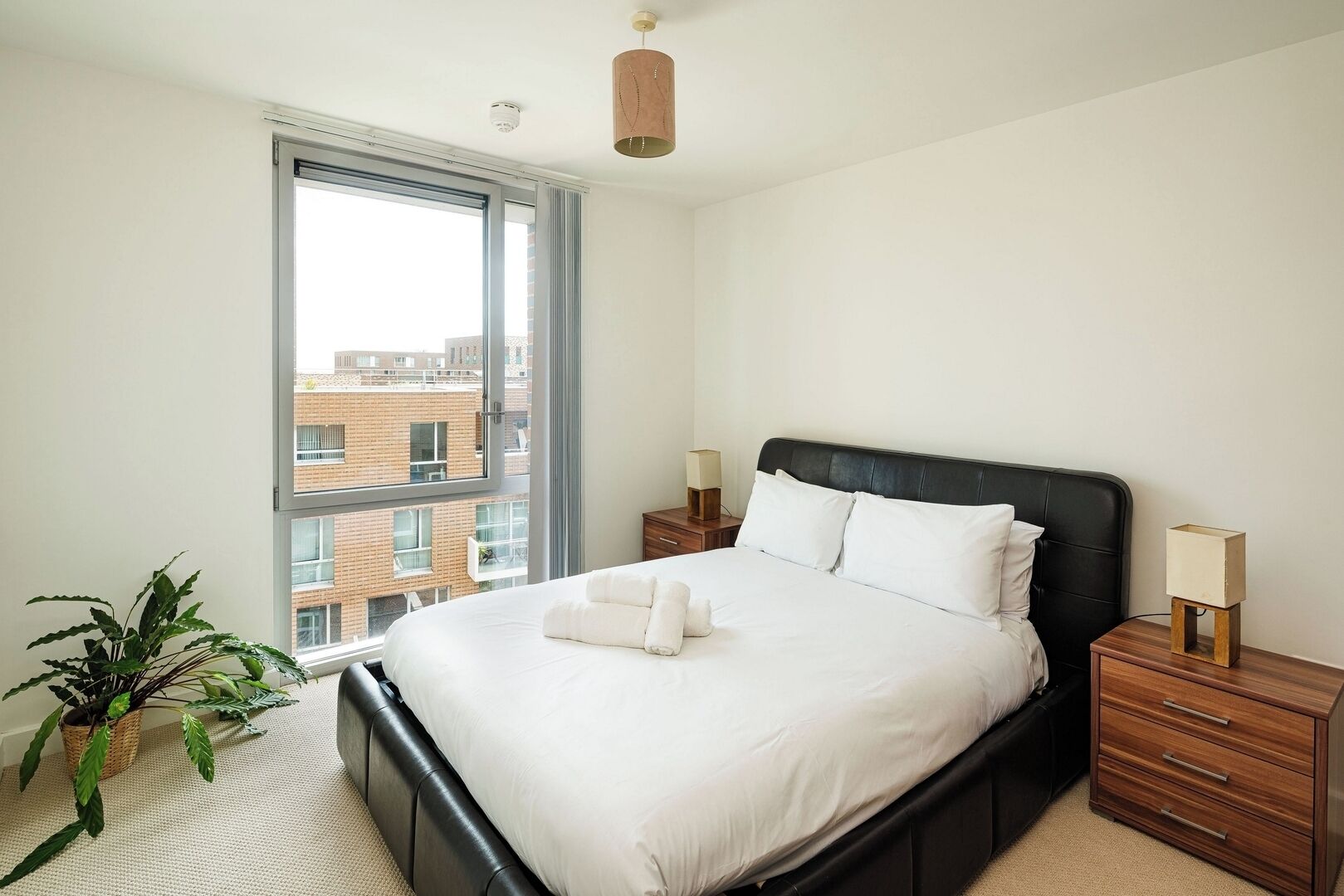 Quarto moderno com uma janela grande, cama com roupa de cama branca, cabeceira escura, mesas de cabeceira com abajures e um vaso de plantas.