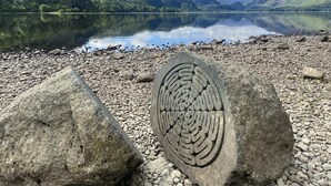 A serene lake with mountains in the background, a pebble beach, and two large rocks with carvings. - 2 Bed in Keswick (oc-95741) (Keswick)