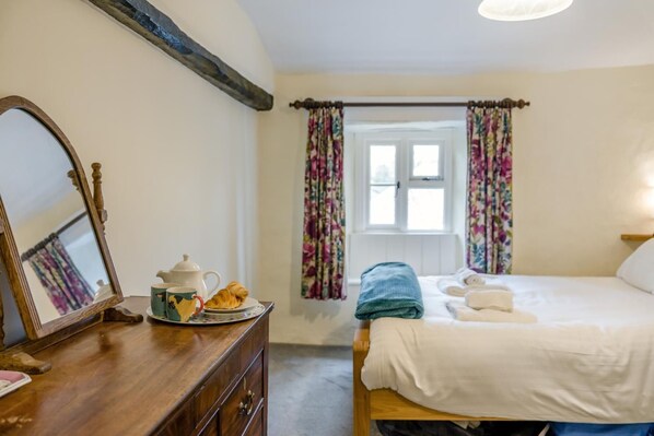 Une chambre avec une commode en bois, un lit recouvert de draps blancs et une fenêtre ornée de rideaux à motifs floraux.