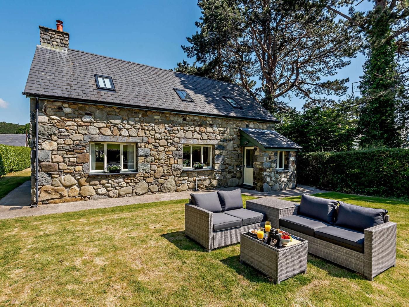 A stone house with a sloped roof, a small porch, and a grassy yard with outdoor furniture.