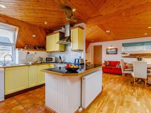 A kitchen with wooden cabinets, a central island, and a dining area with a red sofa and table. - 3 Bed in Bideford (oc-lakco) (Northam)