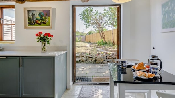 Una cocina con pared de piedra, una mesa de comedor con sillas, una cafetera y un jarrón con flores rojas.
