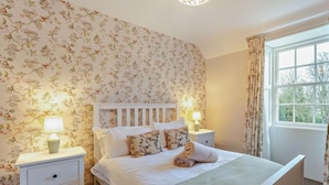 A bedroom with a floral wallpaper, a white bed with a wooden headboard, two bedside tables with lamps, and a window with curtains. - 4 Bed in Hawick (oc-95765) (Teviotdale)